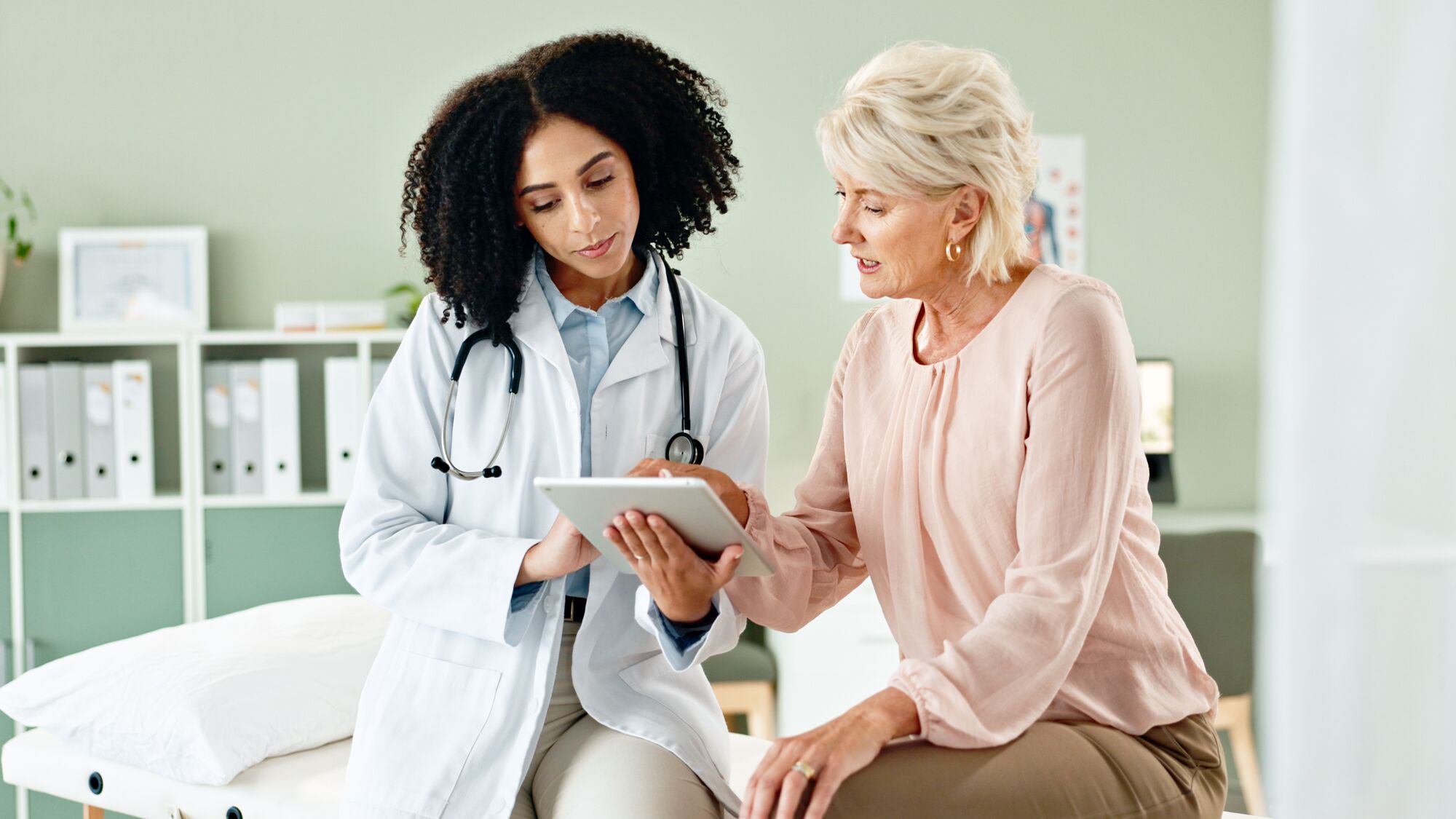 A doctor consulting with a patient pointing at a tablet device in a medical room.
